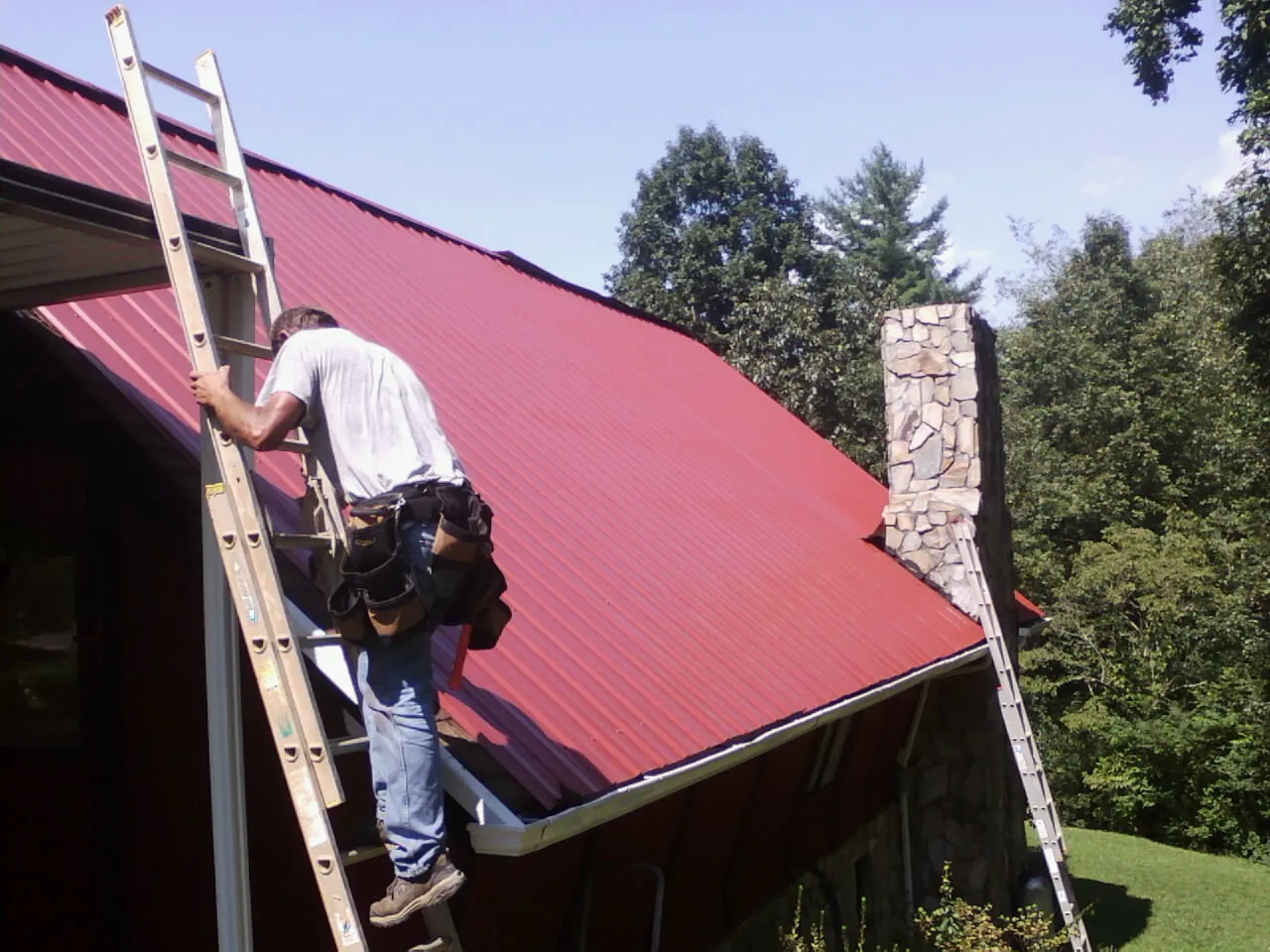 Roofer on a ladder beside a red metal roof and stone chimney — Soffit & Fascia Repair by Roofing Brothers in Olympia